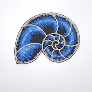 May include: An embroidered patch in the shape of a nautilus shell. The design features vibrant blue thread with black accents and a light tan border outlining the spiral chambers. The embroidery is dense and textured, creating a detailed shell motif.