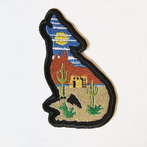 May include: Embroidered patch depicting a coyote silhouette with a desert landscape inside. The coyote is howling at the moon, and the desert scene includes a red rock formation, cacti, and a small adobe house.