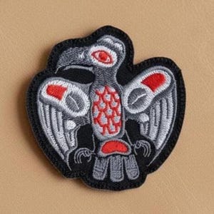 May include: An embroidered patch featuring a stylised bird design in black, grey, and red thread. The bird is depicted with a prominent eye, a patterned chest, and outstretched wings, all outlined with a thick black border on a fabric base.