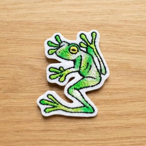 May include: An embroidered patch featuring a green frog with a yellow eye and white border. The frog is depicted in a side profile view with its limbs extended, showcasing detailed stitching in shades of lime and forest green on a white base.