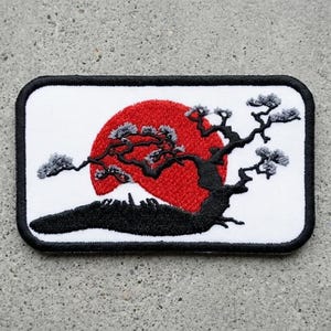 May include: Embroidered patch featuring a Japanese landscape. A red sun rises behind a black silhouette of a mountain and a stylized tree with gray foliage. The patch has a white background and a black border.
