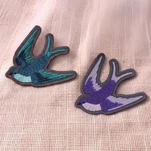 May include: Two embroidered swallow patches. One is teal and green, the other is purple and white. Both have a grey border and are on a textured, off-white background.