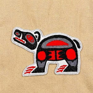 May include: Embroidered patch featuring a stylized bear design in black, red, and gray. The bear has a distinctive tribal art style with a white border, set against a tan fabric background.