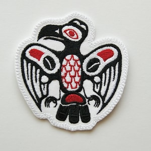 May include: An embroidered patch featuring a stylized bird design in black, white, and red thread. The bird is depicted with wings spread, a prominent beak, and detailed geometric patterns on its chest and wings, all outlined in white stitching.