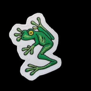 May include: Green embroidered frog patch with yellow eyes on a white background.