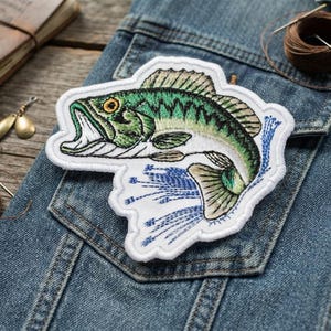 May include: An embroidered patch featuring a green and tan largemouth bass fish leaping from blue water splashes. The fish has a yellow eye and an open mouth, outlined in white thread. The patch is placed on a blue denim fabric surface.