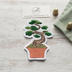 May include: An embroidered patch featuring a bonsai tree with a brown trunk and green foliage in a terracotta-coloured pot. The patch is white-bordered and accompanied by a card labelled Artisan Quality Embroidered Patch - 7.6 cm H and two gold thimbles.