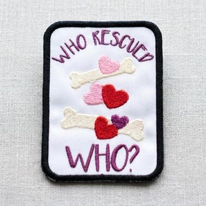 May include: A rectangular embroidered patch with a white background and a black border. The text "WHO RESCUED WHO?" is stitched in purple. The design features two cream-coloured bones, a light pink heart, a red heart, and a small purple heart.