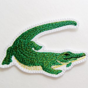 Peut inclure: Un écusson brodé représentant un crocodile vert avec une queue courbée, détaillé avec des points de broderie vert foncé pour le corps et des points jaune clair le long du ventre, fini avec une bordure blanche cousue tout autour.