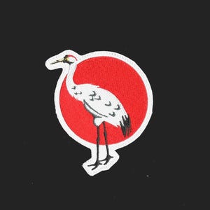 May include: White embroidered crane with black details standing on a red circle with a white outline.