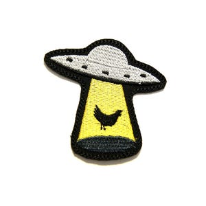 May include: A black and yellow embroidered patch depicting a UFO with a black silhouette of a chicken inside. The UFO is flying over a yellow beam of light.