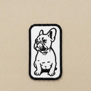 May include: A rectangular embroidered patch featuring a black and white line art illustration of a French Bulldog. The patch has a white background with a thick black border and is centred on a solid light beige fabric surface.