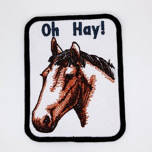 May include: Embroidered patch with a horse head in brown and white, and the text "Oh Hay!" in navy blue. The patch has a black border and is on a white background. Ideal for adding to clothing or accessories, this patch is a fun, equestrian-themed item.
