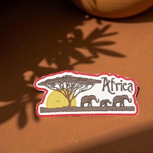 Peut inclure: Écusson brodé avec une bordure rouge représentant une scène de savane africaine. Le motif comprend un arbre, un soleil jaune et trois éléphants. Le mot "Africa" est écrit dans une police stylisée.
