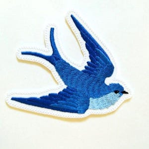 May include: A blue embroidered swallow bird patch with white stitching around the edges.