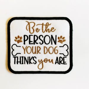 May include: Embroidered patch with a white background and a black border. The patch features the text "Be the PERSON YOUR DOG THINKS you ARE" in brown and black, along with paw prints and a bone graphic.