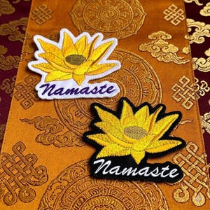 May include: Two embroidered patches featuring a yellow lotus flower with a textured center. One patch has a white border and the word Namaste in purple script, while the other has a black border and the word Namaste in white script.
