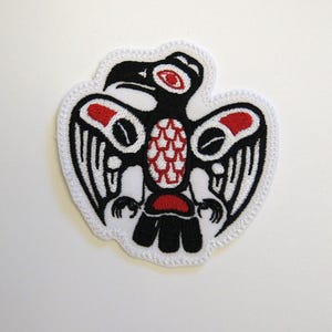 May include: An embroidered patch featuring a stylized bird design in black, white, and red thread. The bird is depicted with a prominent beak, an oval eye, and detailed wing patterns, all outlined with a white border on a white fabric base.