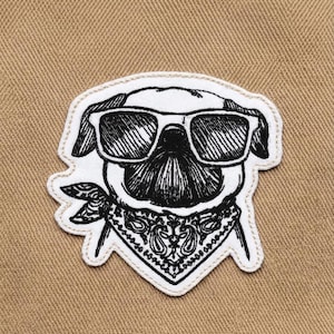 Op de afbeelding: Geborduurde patch met een mopshond met een zonnebril en een bandana. De patch is wit met zwarte details, waaronder het gezicht van de hond, de zonnebril en het bandanamotief. De achtergrond is een bruinachtige stof.