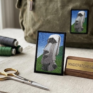 May include: Embroidered patch featuring a Moai statue against a blue sky and green hills. The patch is framed in black and displayed with spools of thread, scissors, and a wooden sign that reads MAGICPATCHES&MORE.