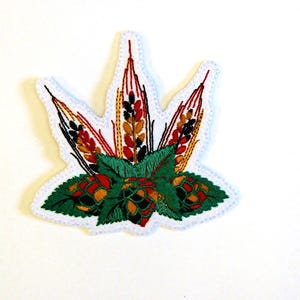 May include: A white fabric patch shaped like a leaf, featuring embroidered details of golden wheat stalks and green hop cones with dark green leaves. The design is outlined with a white stitched border, creating a textured and colourful botanical motif.