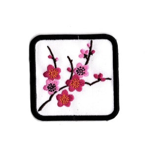 Könnte beinhalten: Gestickter Aufnäher mit einem weißen Quadrat und einem schwarzen Rand. Er zeigt einen Zweig mit rosa und dunkelrosa Kirschblüten mit gelben Zentren. Das Design ist detailliert und eignet sich für Kleidung oder Accessoires.