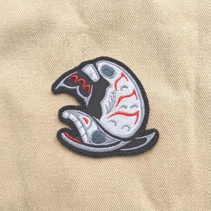 May include: Embroidered patch with a black outline and a stylized design. The patch has a grey background with red and blue accents. The design appears to be a stylized creature or object. The patch is on a tan fabric background.