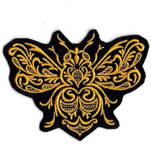Peut inclure: Écusson brodé représentant une abeille stylisée. Le patch a un fond noir avec une broderie dorée complexe. Les ailes et le corps de l'abeille sont ornés de motifs tourbillonnants.