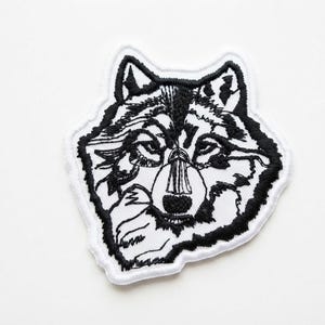 May include: An embroidered patch featuring a stylised wolf head design. The artwork is rendered in black thread against a white background, with a thick white border outlining the shape of the wolf's head, ears, and fur, creating a high-contrast look.