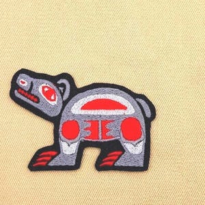 May include: Embroidered patch featuring a stylized bear design in gray, red, and white. The bear has a black outline and is depicted in a traditional Native American art style. The patch is likely for clothing or accessories.