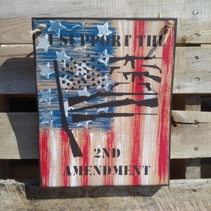 May include: Wooden sign with an American flag design and the text "I Support the 2nd Amendment". The flag is painted in red, white, and blue with a black silhouette of guns.