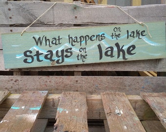 May include: A rectangular wooden sign with the phrase "What happens on the lake stays on the lake" in black lettering. The sign is painted light green with white and blue accents and is suspended by a thin rope.