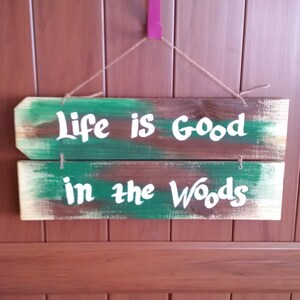 May include: A wooden sign with green and brown paint, hanging on a brown wooden door. The sign reads "Life is Good in the Woods" in white paint.