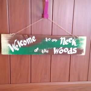 May include: A wooden sign with the words "Welcome to our Neck of the Woods" painted in white on a green and brown background. The sign is hanging from a string.