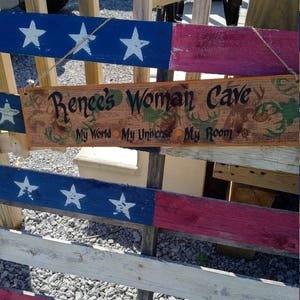 May include: A wooden sign with the text "Renee's Woman Cave" and the words "My World, My Universe, My Room" painted on it. The sign is hanging on a wooden fence painted with red, white, and blue stripes.