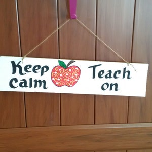 May include: A white wooden sign with the text "Keep Calm Teach On" and a red apple with yellow and white swirls painted on it.