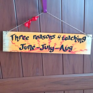 May include: A wooden sign with orange and red paint and black text that reads "Three reasons 4 teaching June-July-Aug". The sign is hanging from a string on a brown wooden door.