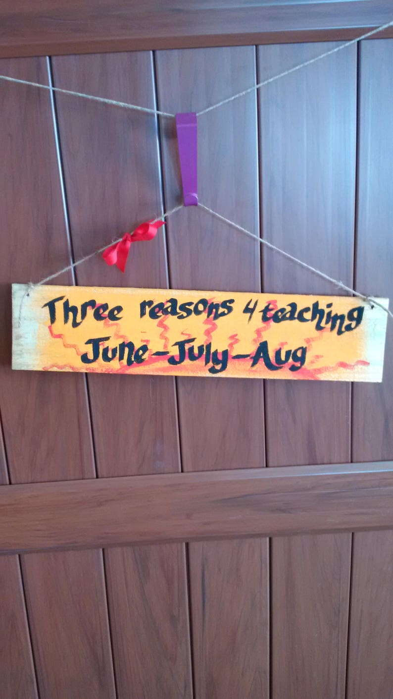 May include: A wooden sign with orange and red paint and black text that reads "Three reasons 4 teaching June-July-Aug". The sign is hanging from a string on a brown wooden door.