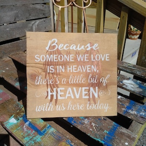 May include: A wooden sign with white painted text that reads "Because someone we love is in heaven, there's a little bit of heaven with us here today."