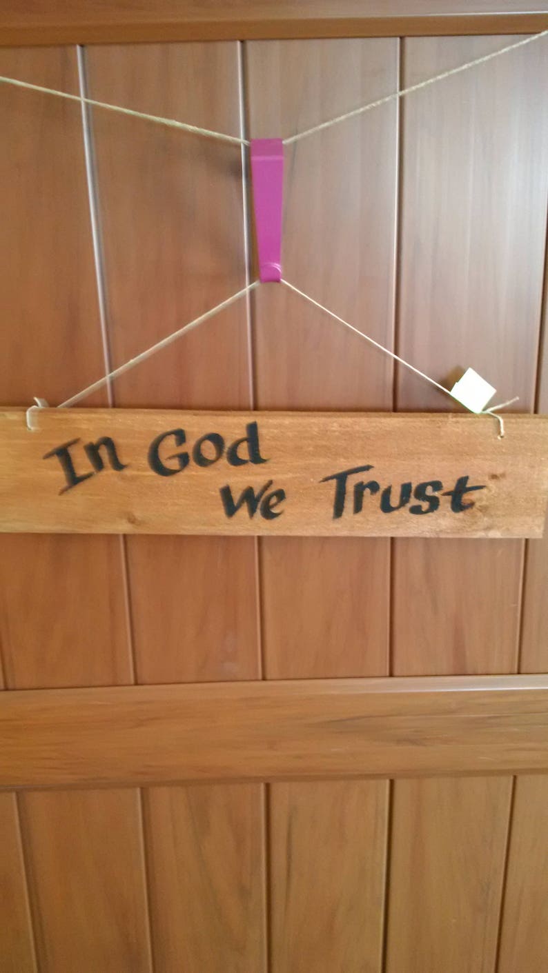 May include: A wooden sign with the text "In God We Trust" hanging on a wooden door. The sign is attached to the door with twine and a pink plastic hook.