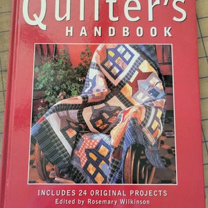 May include: The Quilter's Handbook book cover features a red background with the title in white text. The cover image shows a colorful quilt draped over a wooden chair. The text "Includes 24 Original Projects" and "Edited by Rosemary Wilkinson" is printed below the image.