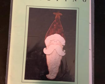 May include: A quilting pattern titled "The Santa Claus Wall Hanging" by Curtis Boehringer. The pattern features a Santa Claus design with a red hat, white beard, and a star on top. The pattern is in a plastic sleeve.