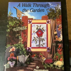 May include: A book cover titled "A Walk Through the Garden" featuring a colorful quilt design with a birdhouse and garden scene. The cover includes the word "Vegetables" and is by "Piecemakers".