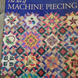 May include: The Art of Machine Piecing book cover features a colorful quilt design with various geometric patterns and a blue background. The title "The Art of Machine Piecing" is written in gold lettering, and the author's name, "Sally Collins", is written in white lettering.