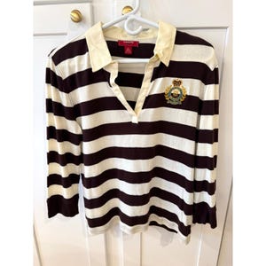 Puede incluir: Una camisa de manga larga a rayas en marrón y crema. La camisa presenta un cuello color crema y un escudo en el pecho. La marca "CHAPS" es visible en la etiqueta. La camisa está colgada en una percha blanca.
