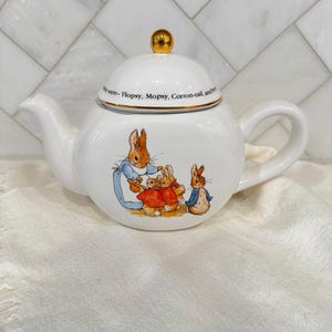 May include: White ceramic teapot with a gold-coloured knob on the lid. The teapot features a colourful illustration of rabbits in clothing. The text "were- Flopsy, Mopsy, Cotton-tail, and Peter" is printed on the rim.