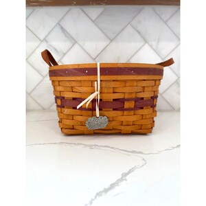 May include: A rectangular woven basket in natural wood tones with burgundy stripes. It features leather handles and a decorative metal charm suspended from a white ribbon. The basket measures approximately 25 cm long, 20 cm wide, and 15 cm tall.