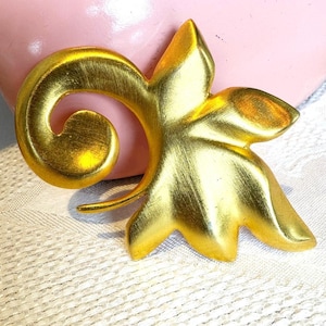 May include: A gold-toned metal brooch with a leaf and swirl design. The brooch has a textured surface and is set against a pink background. The brooch is a decorative accessory.
