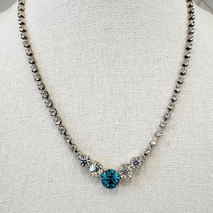 May include: A silver-toned necklace with clear and teal-colored rhinestones. The necklace features a central teal-colored rhinestone surrounded by smaller clear rhinestones. The chain is composed of small, clear rhinestones.
