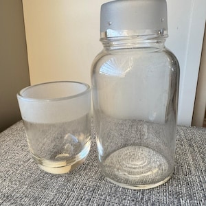 Może przedstawiać: Przezroczysta szklana butelka z matową pokrywką i mały, przezroczysty szklany kubek. Butelka ma zaokrąglony kształt i szeroki otwór. Kubek jest krótki i szeroki, z matowym paskiem na górze. Oba są puste.